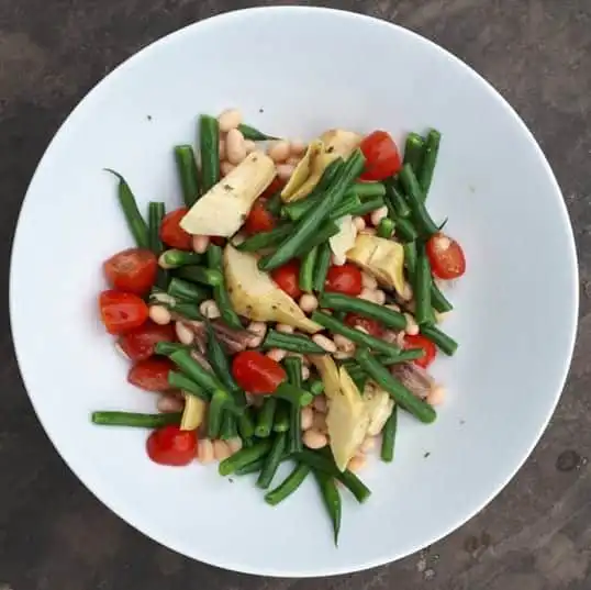 Round white bowl holding salad made of cut green beans, artichokes, tomatoes, anchovies and white beans