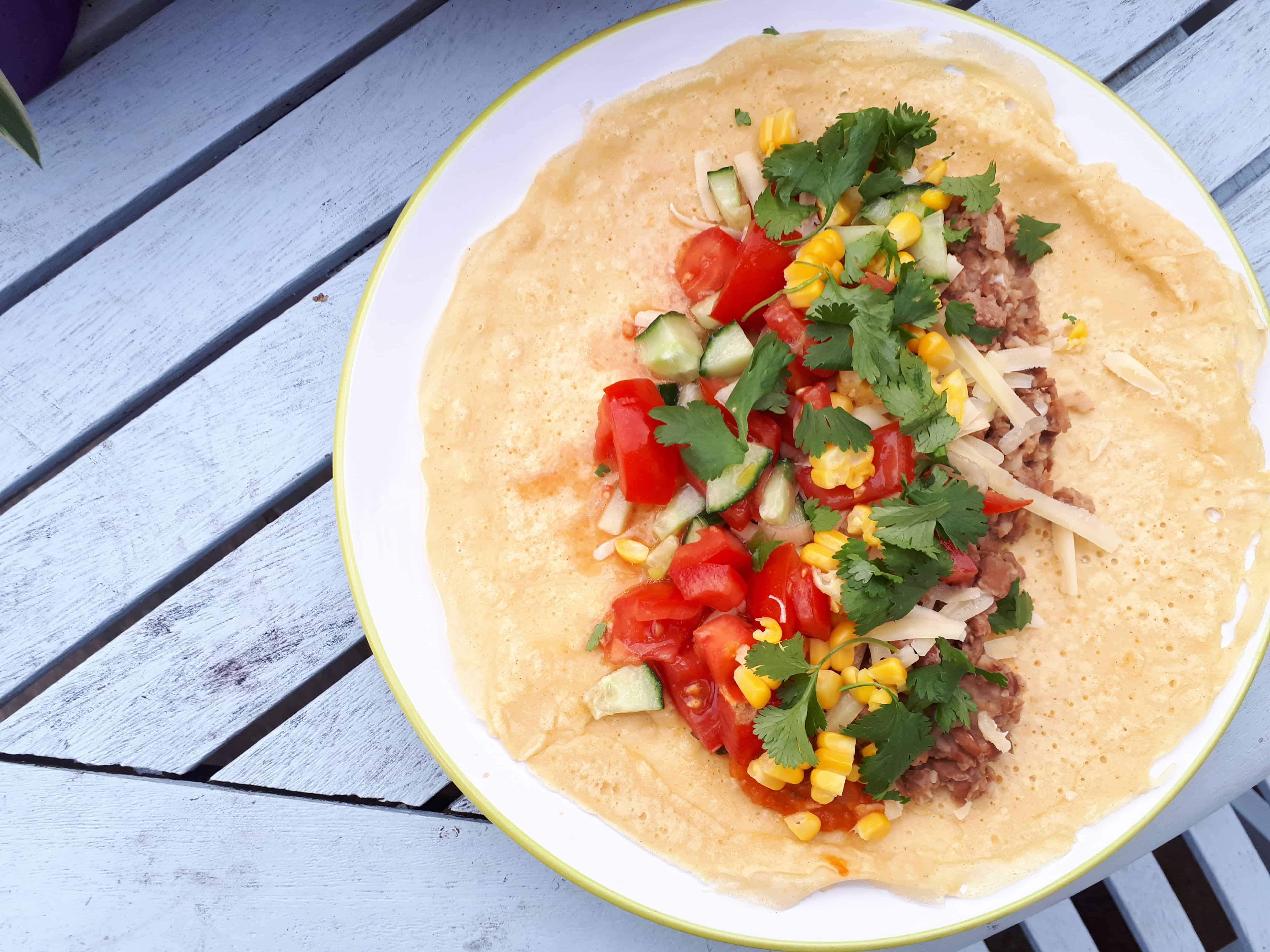 gram flour wrap with tomatoes, sweetcorn, refried beans, coriander and cheese, against a light blue table