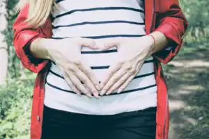 white woman making a heart shape with her hands on her belly. wearing a white and blue striped top and red jacket