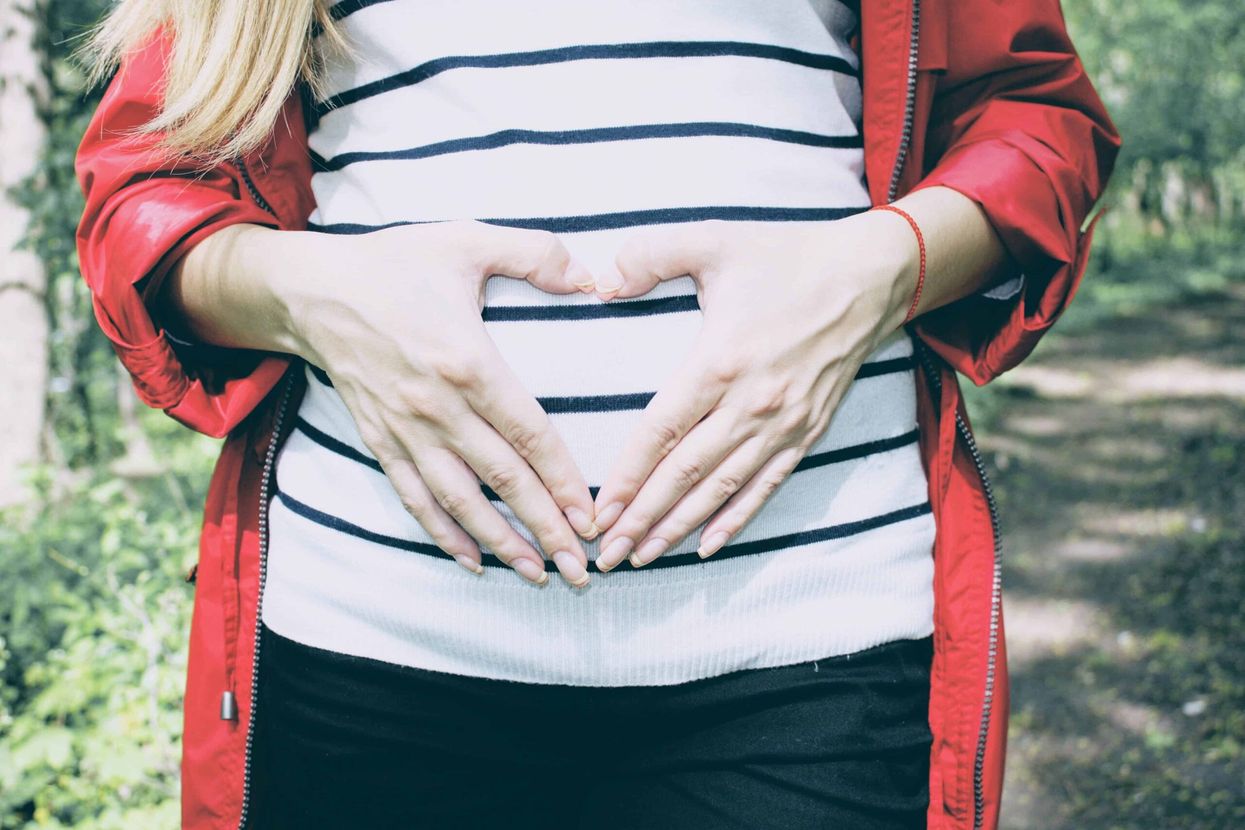 white woman making a heart shape with her hands on her belly. wearing a white and blue striped top and red jacket