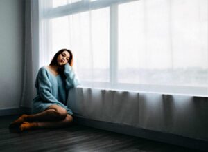 woman sitting on the floor looking sad in an empty room next to big window and a white curtain
