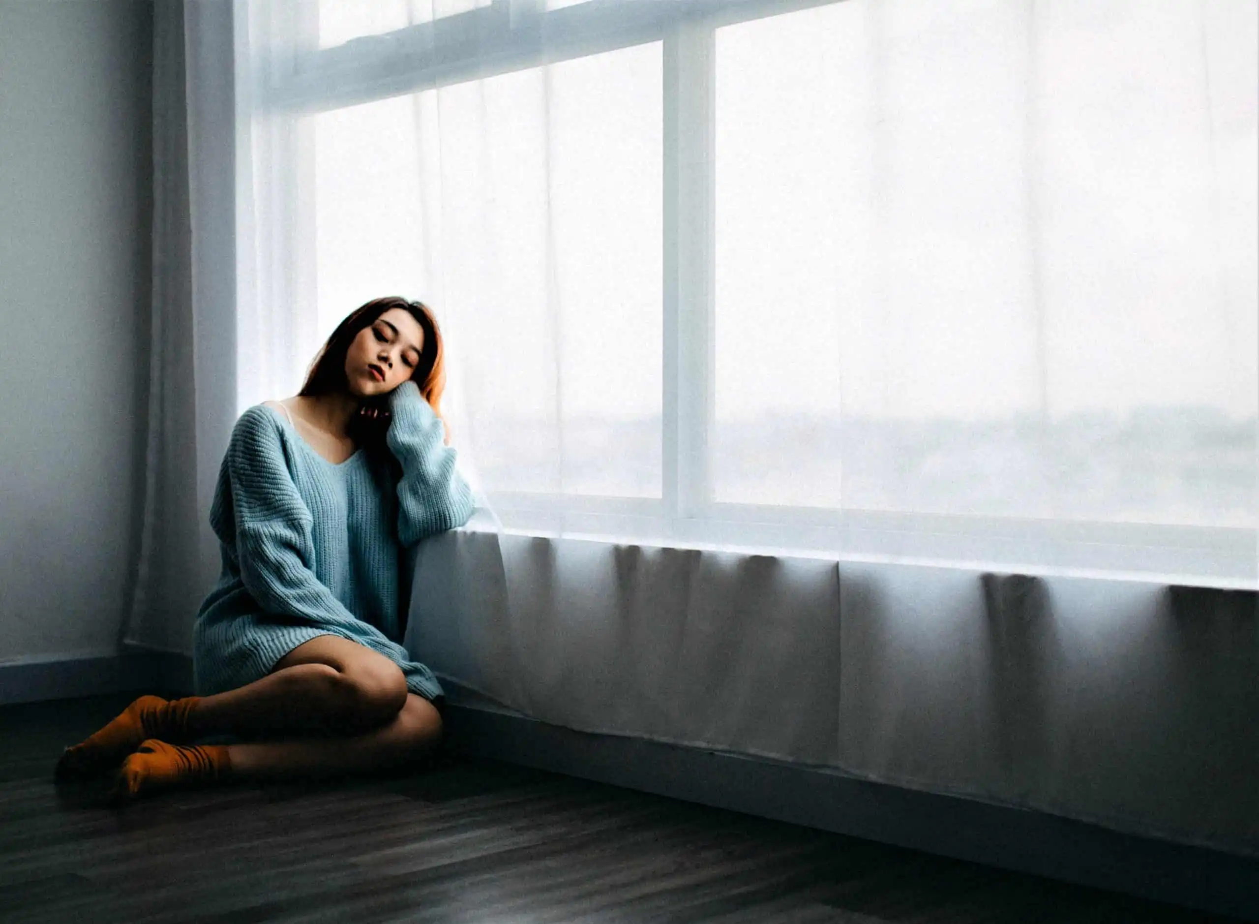 woman sitting on the floor looking sad in an empty room next to big window and a white curtain