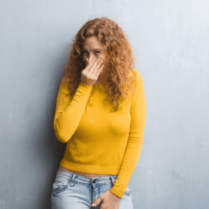 white woman with curly hair, wearing a yellow jumper and jeans holding her nose