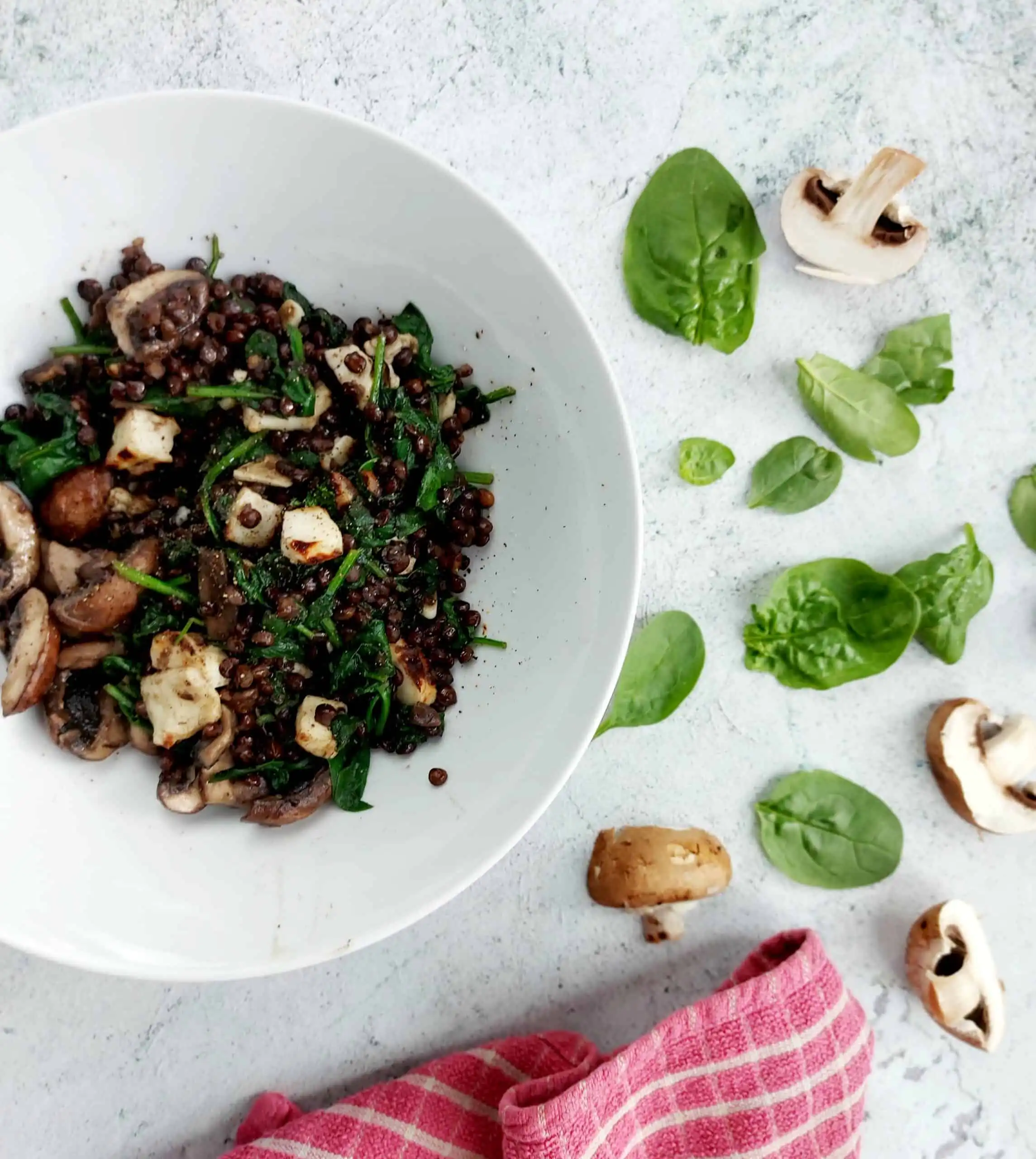 white bowl containing lentil, mushroom, spinach and halloumi salad against pale blue background, raw chopped mushrooms and spinach scattered around
