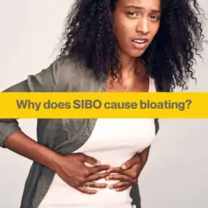 woman clutching her stomach looking pained. She wears white t-shrt with grey cardigan, has black skin and black curly hair.
