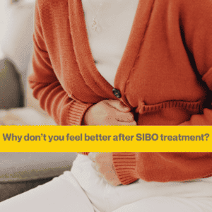 Woman's torso close up. She is wearing an amber cardigan and white T shirt, white trousers. She is clutching her stomach. The words Why you don't feel better after SIBO treatment.