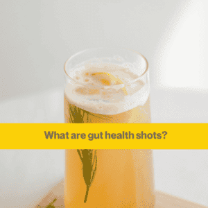Glass containing some orangey yellow liquid and a sprig of rosemary. The background is a white wall with sunlight in the coner. 