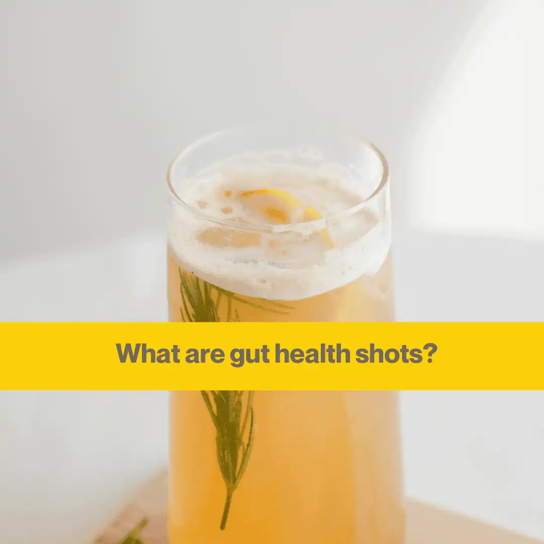 Glass containing some orangey yellow liquid and a sprig of rosemary. The background is a white wall with sunlight in the coner.