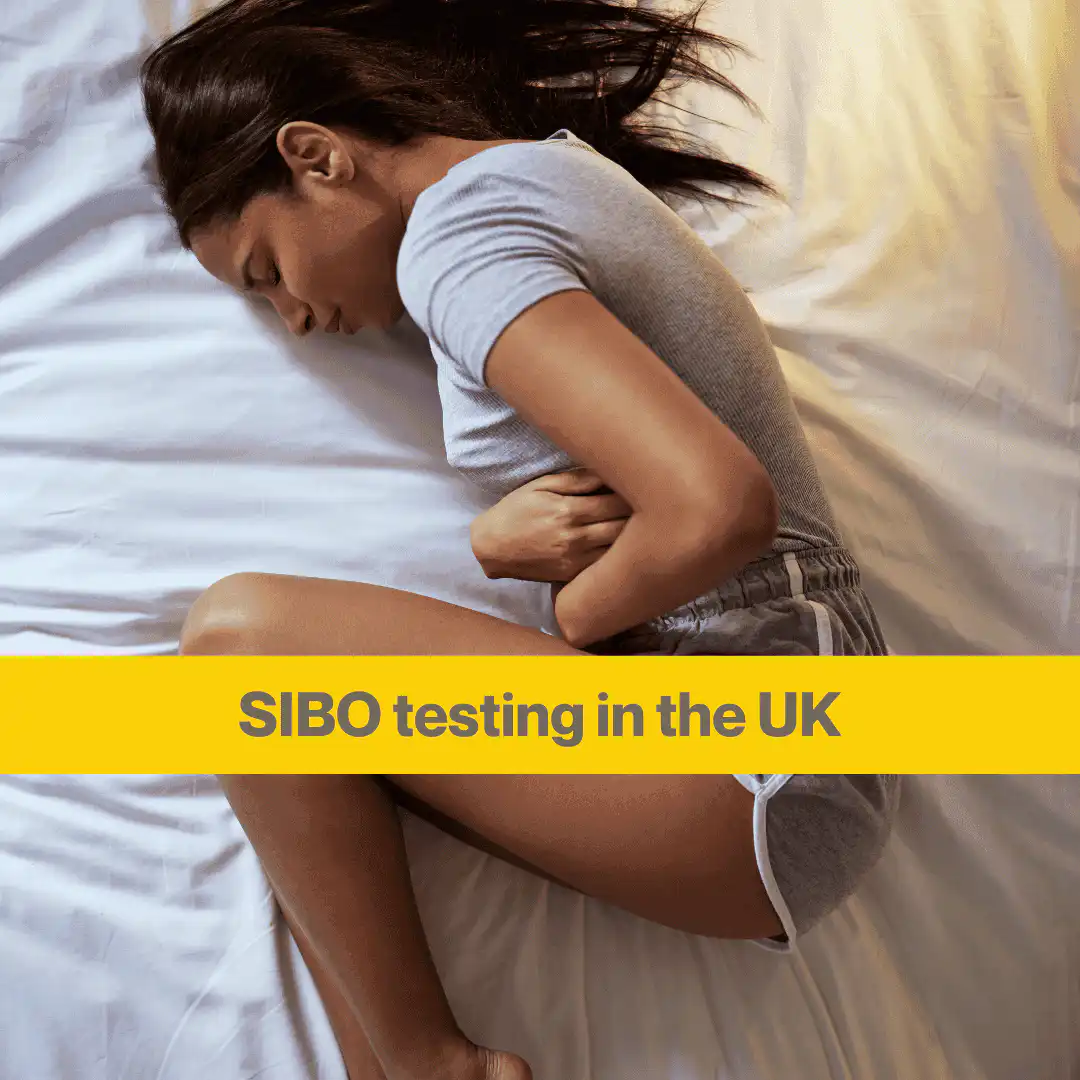 woman with long dark hair lying on a white bed. She's in a foetal position and holding her stomach. Words SIBO testing in the UK