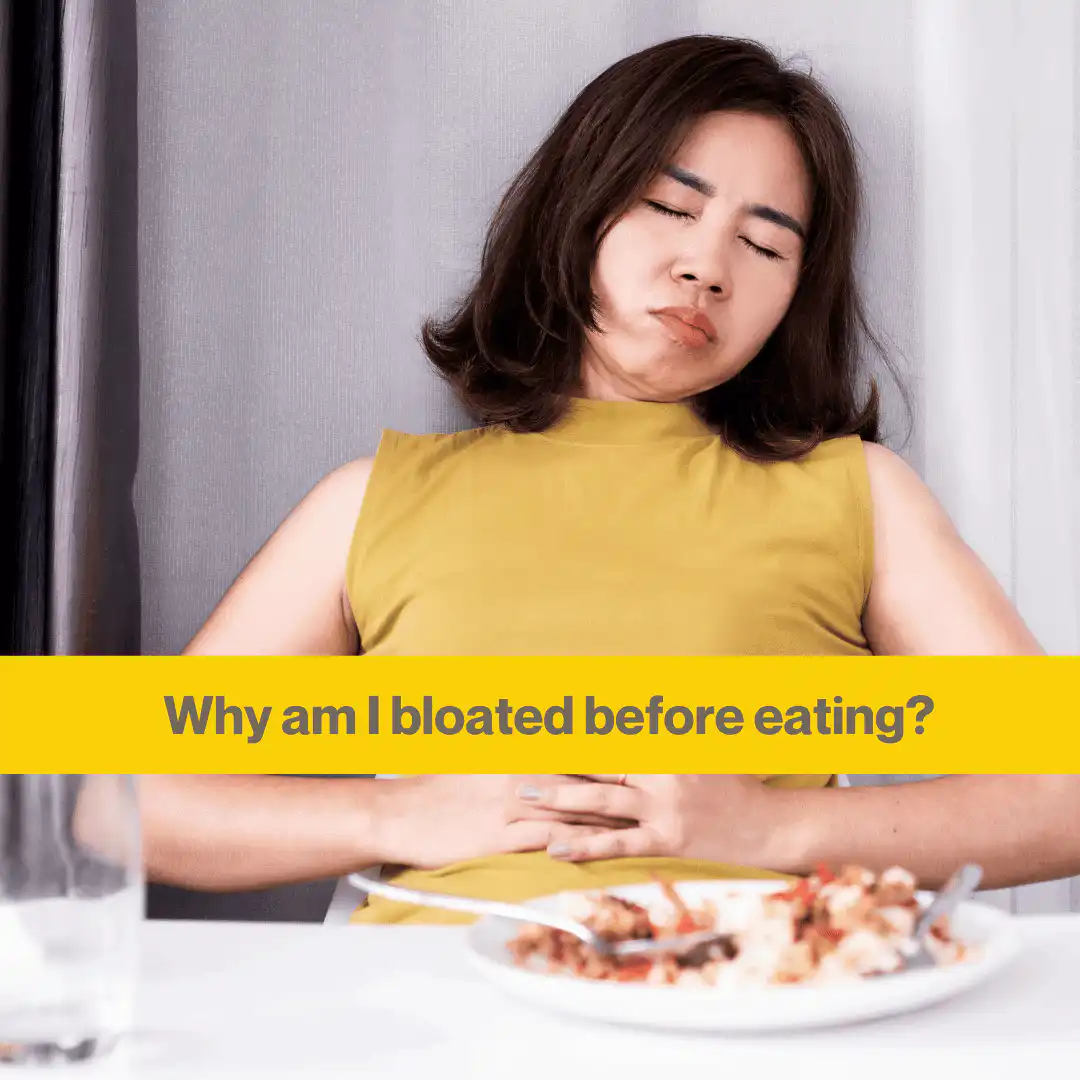 woman with black hair and a yellow sleeveless high necked top leans back from a table with eyes closed holding stomach.