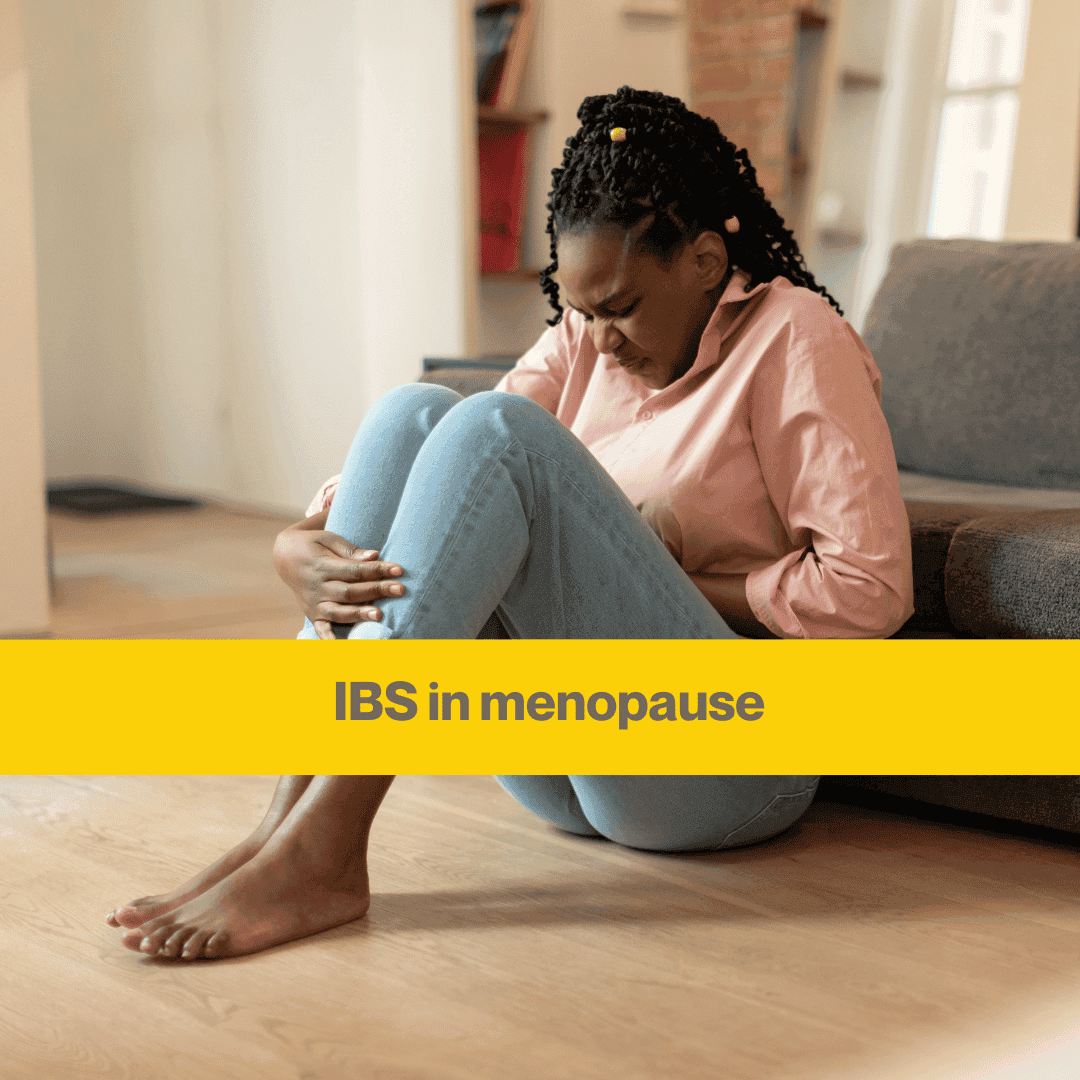 Woman sitting on floor next to a grey couch. She's wearing tight skinny blue trousers and pink top, knees up clutching stomach. Words IBS in menopause